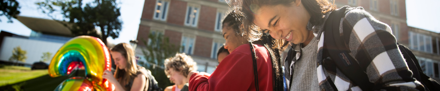 Students at an outdoor event. The student closest to the camera has brown, shoulder length wavy hair and is smiling while looking down.