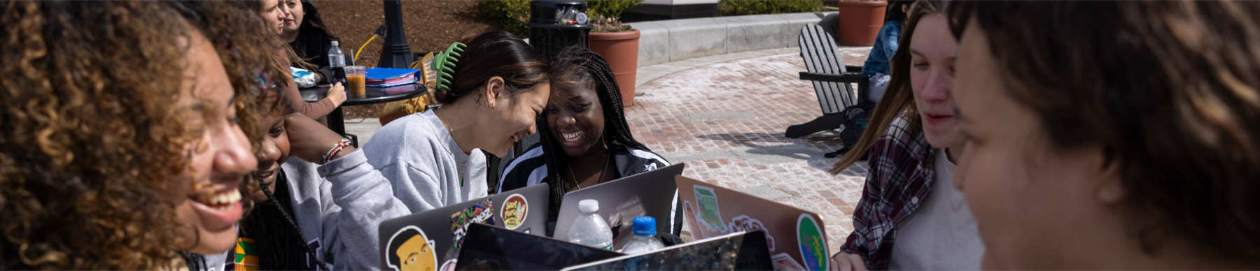 A group of diverse students outside on a sunny day studying. There about 6 students sitting around a round table with laptops.
