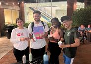 Four students standing behind a craft table, smiling. Two students are holding an ice cream cup. Another is holding a bottle of paint. There are various paint bottled with different colored paint on the table.