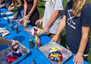 Three students standing at a table with a blue tablecloth, smiling on a sunny day.