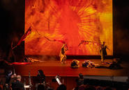 Dancers on-stage against a red backgroung; the orchestra in the foreground.