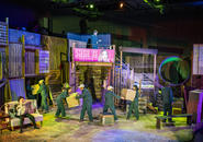 Busy workers in the Shen Te factory; Shui Ta ignores the activity and reads newspaper.  Sun is in charge.