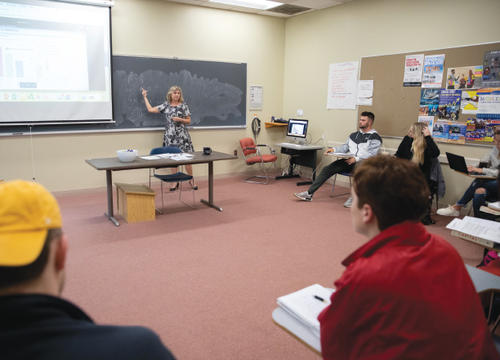 Kolleen Rask points at a blackboard in a bright classroom full of students