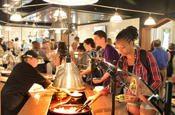 Crowd of students helping themselves in the servery of Kimball Dining Hall, as a worker behind the servery replenishes dishware.