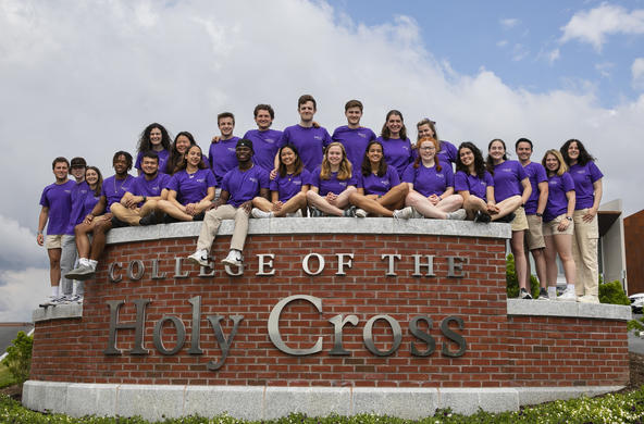 Lots of students wearing purple Holy Cross shirts sitting on the College of the Holy Cross wall.