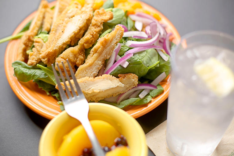 Plate of grilled chicken salad, ice water with lemon wedge, and a small bowl of fruit with a fork resting on it.