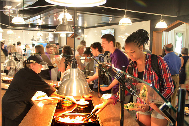 Crowd of students helping themselves in the servery of Kimball Dining Hall, as a worker behind the servery replenishes dishware. 