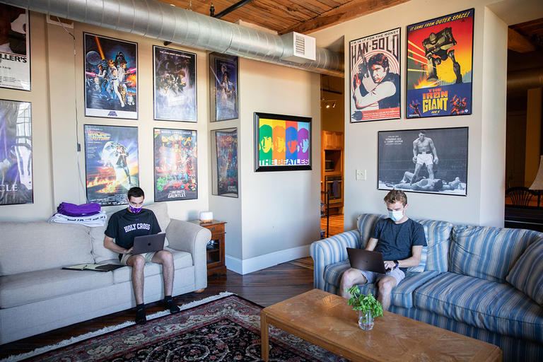 two students sitting on separate couches and working on laptops in their apartment room with posters decorating the walls