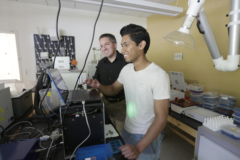 student working on a laptop in a science lab while his faculty mentor looks on