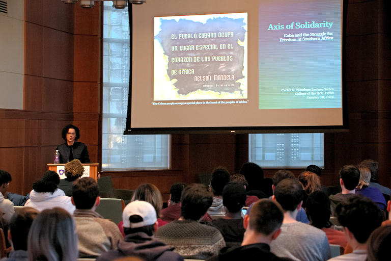 person speaking to a group of students a presentation is on the screen there are windows in the background