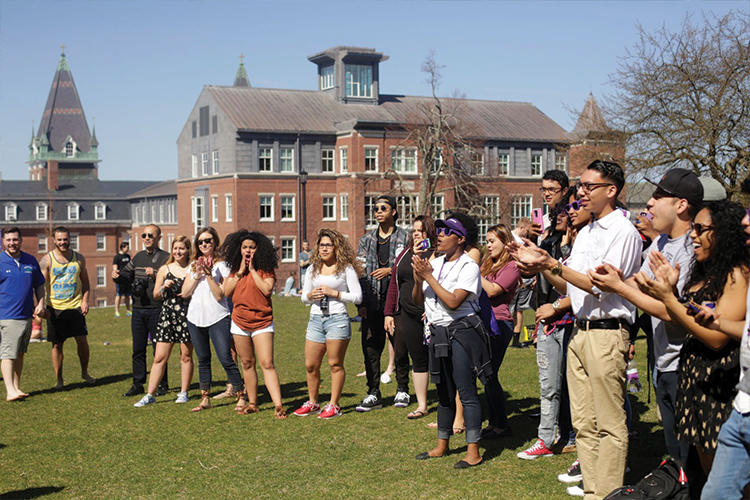 Outdoor campus view with large stone building in the background.  Congregation of students in the foreground.