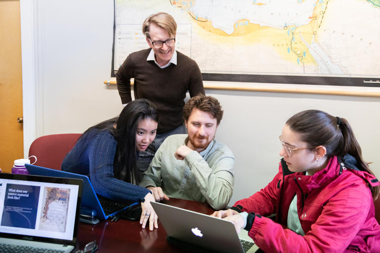 Students and instructor gathered in a classroom around their laptops working together.