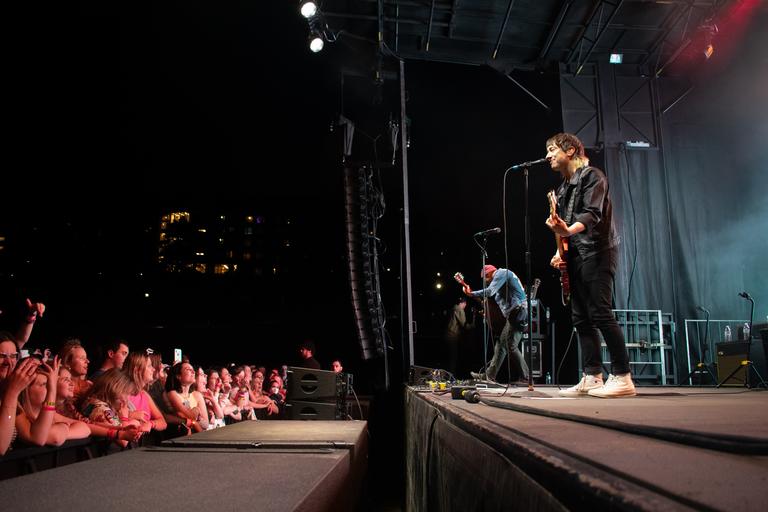 Photo of a ban on stage in the evening with fans on the ground below having a great time. The band members in the photo include a person wearing all black, playing guitar and singing and another guitarist in a blue shirt and red hat.