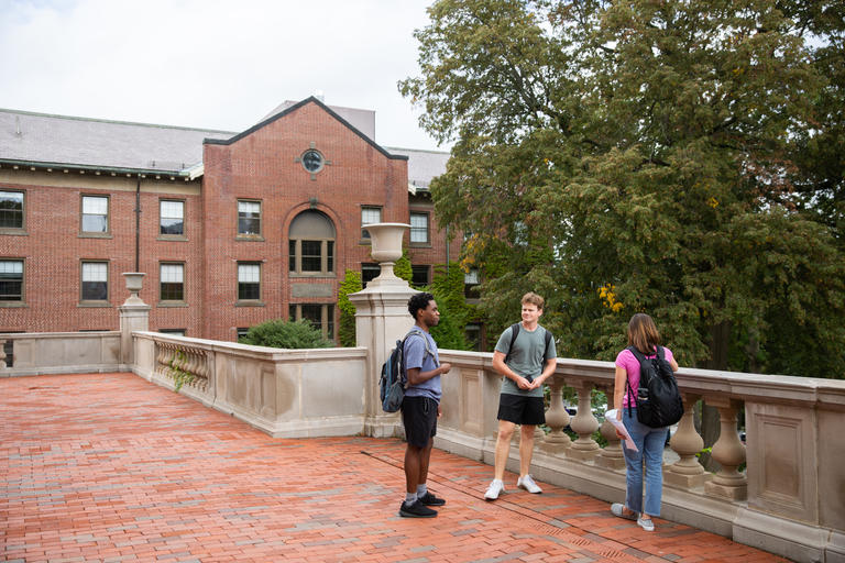 3 Students on campus talking to each other while standing on a brick walkway in front of Dinand Library