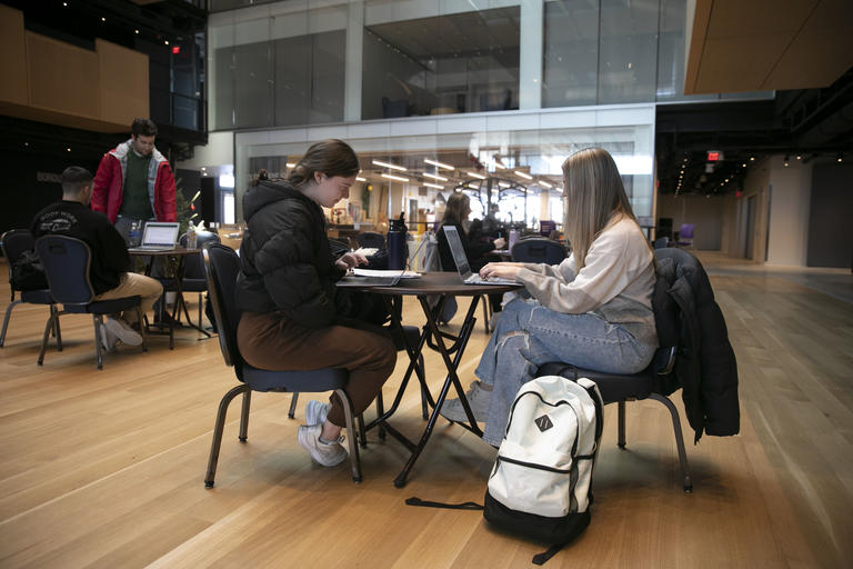 In focus are two students sitting at a round table in a study hall. They are each on their laptops.