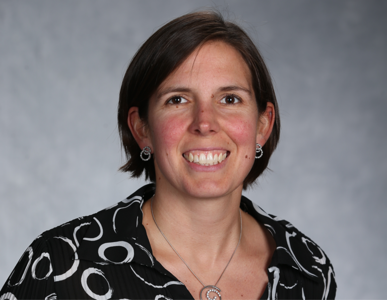 A portrait photo of Jamie Shannon. Jamie has chin length brown hair, parted on teh side. She has brow eyes and is wearing earrings and a black shirt button down shirt with white swirls. She is also wearing a silver chain necklace with some sort of pendant.