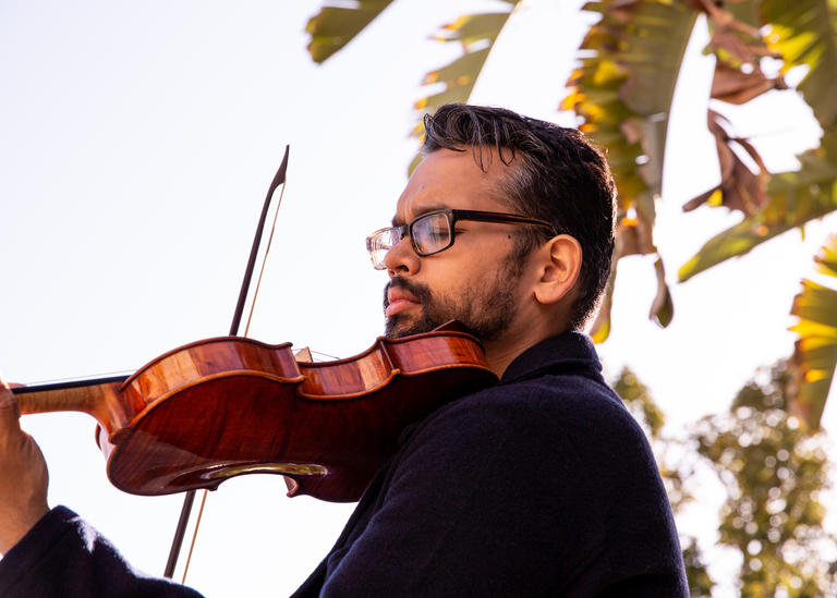 A man playing a violin outside with trees in the background.