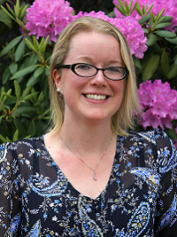woman with light hair pattered shirt in blue, white and green flowers and pink flowers behind her