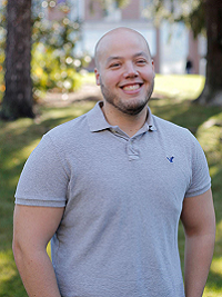 close up picture of man in gray shirt with building and trees behind him