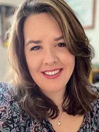 close up of a women with long light brown hair wearing a dark pattered dress with a blurry back ground.  She is smiling at the camera