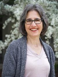 woman outside with green bush behind her light colored shirt and gray sweater, short hair and glasses smiling at the camera