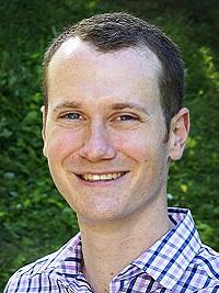 close up of male faculty member wearing a plaid shirt light blue, short brown hair smiling at the camera with trees in the background