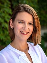 close up of a young woman, hair swept to the side wearing a white shirt smiling at the camera with a dark green background