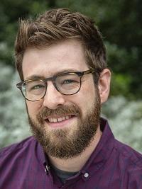 close up of a man with short dark hair a beard and mustache wearing glasses and a plaid shirt smiling at the camera