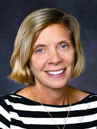 close up picture of a woman smiling at the camera, short blond hair, dark background wearing a black and white striped sweater.