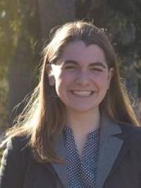 Margaret, who wears a grey coat over a patterned shirt, stands outside in front of pine trees. Margaret has straight brown hair and smiles.